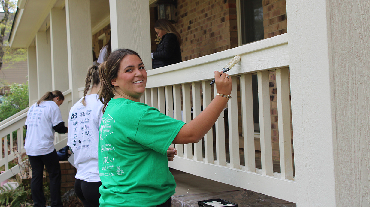 A young woman wearing a green T-shirt smiles as she helps paint a porch railing as two other young women work nearby.