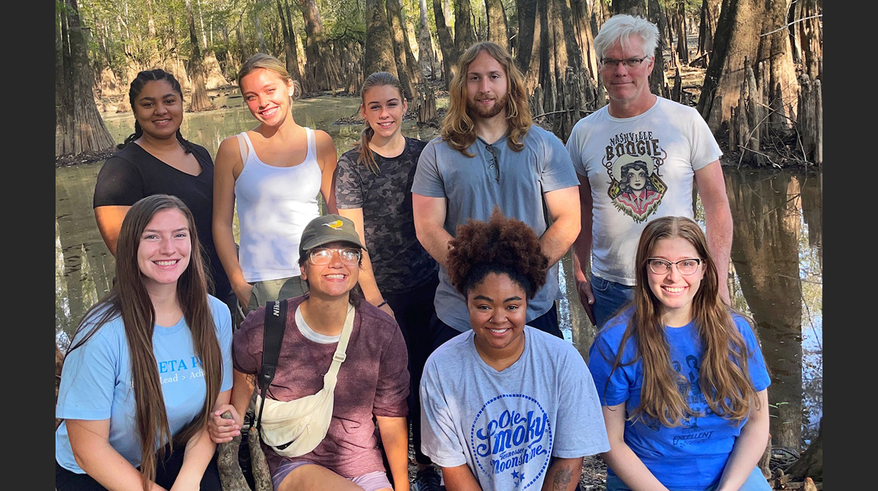 A group of young people stands in front of a swampy area lined with cypress trees.