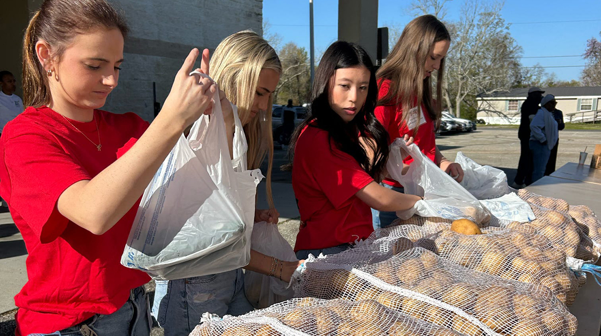 Four young women sort potatoes into white plastic grocery bags while standing at an outdoor table covered with mesh bags of produce.