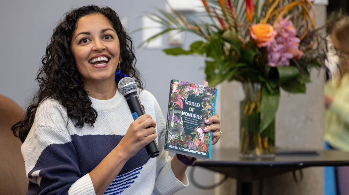 A woman holds a microphone and a book while sitting on a stage.