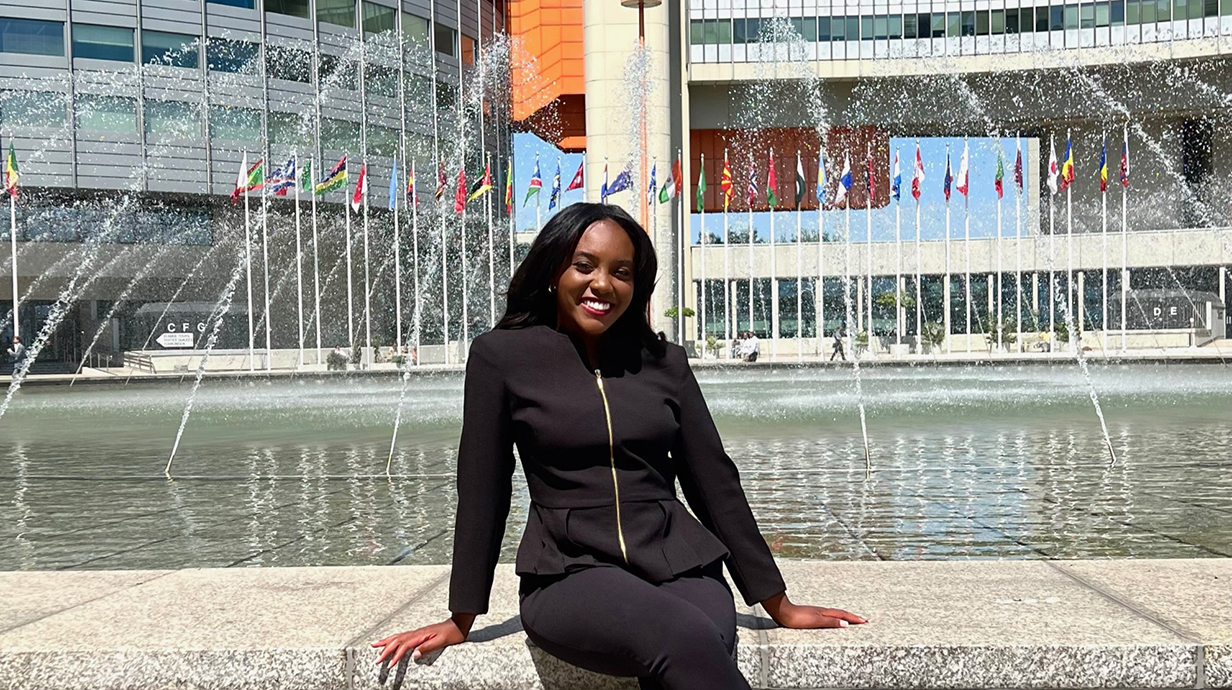 A young woman sits on a fountain in front of modern concrete-and-glass buildings in a downtown area.