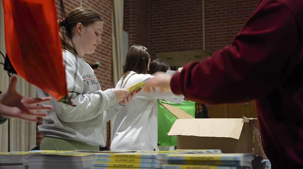 A young woman wearing a gray sweatshirt looks at a book while others pack books into boxes.
