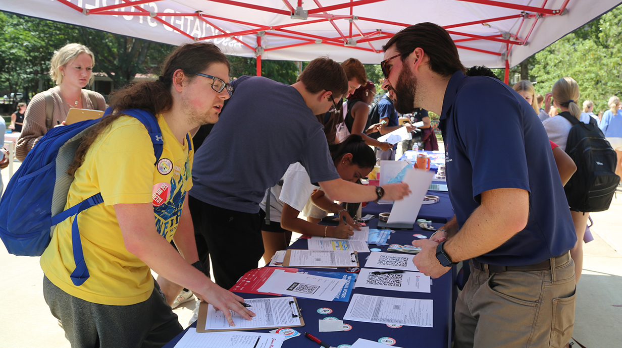 Two men standing on opposite sides of a blue table underneath a pop-up tent talk while other people work with papers on the table on a sunny day.