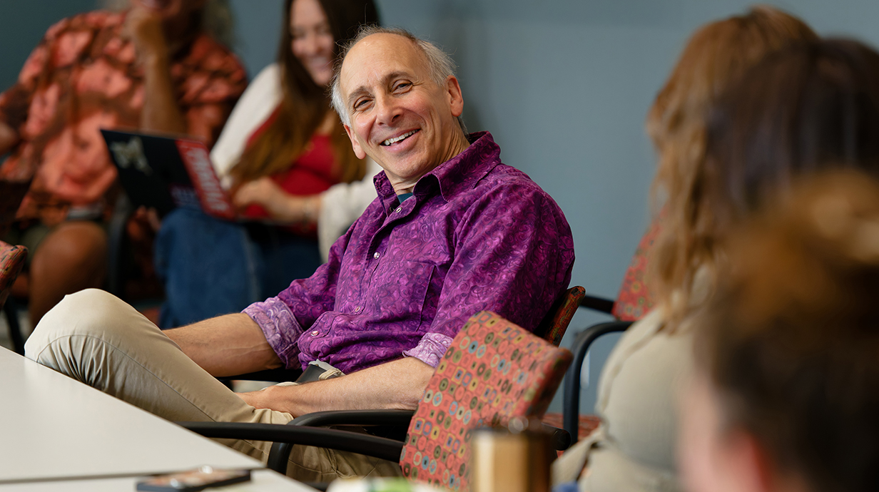 A man wearing a purple shirt and seated at a table smiles and talks to others at the table.