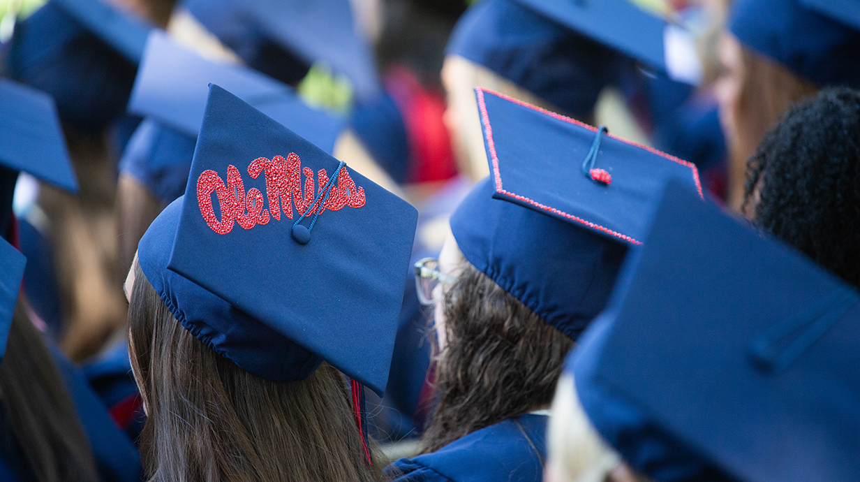 Rows of people dressed in blue graduation robes and mortar boards -- one decorated with 'Ole Miss' in red script -- sit in a ceremony.