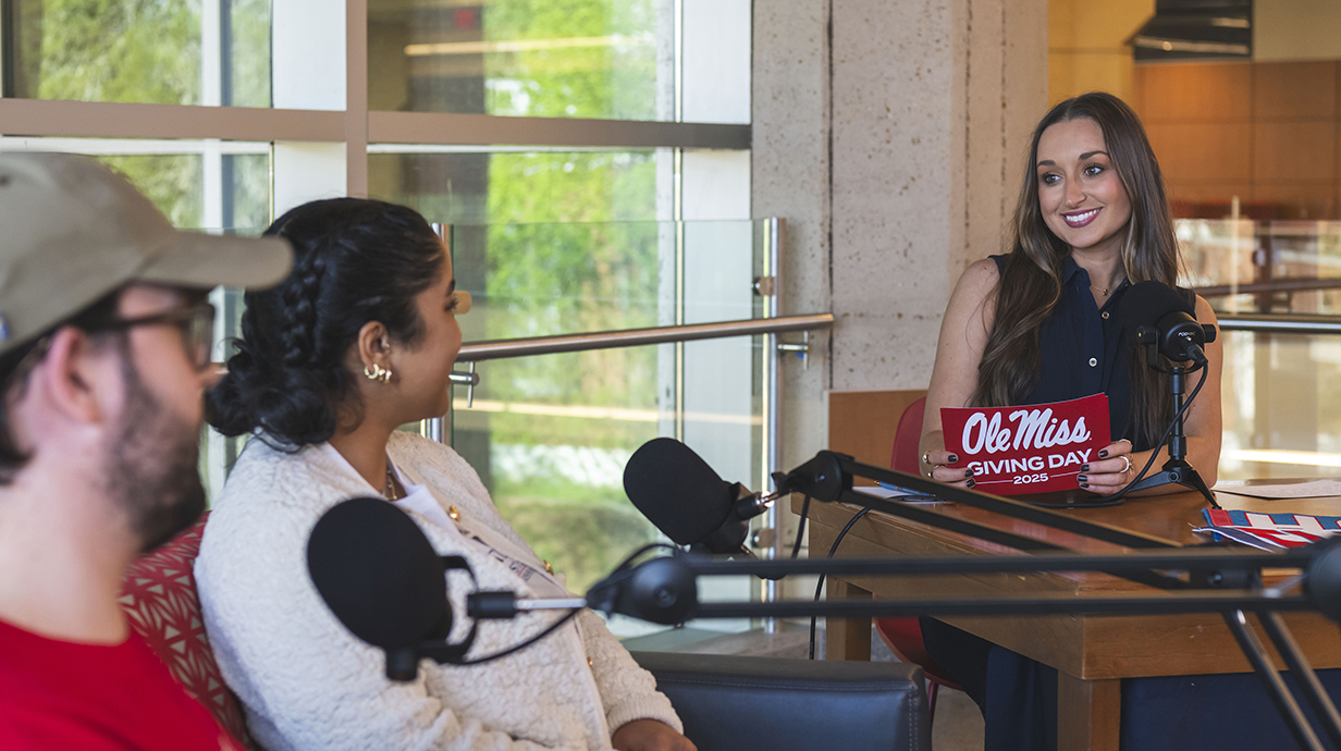 A young woman holding a stack of red cards reading 'Ole Miss Giving Day 2025' interviews two young people sitting behind a microphone stand nearby.