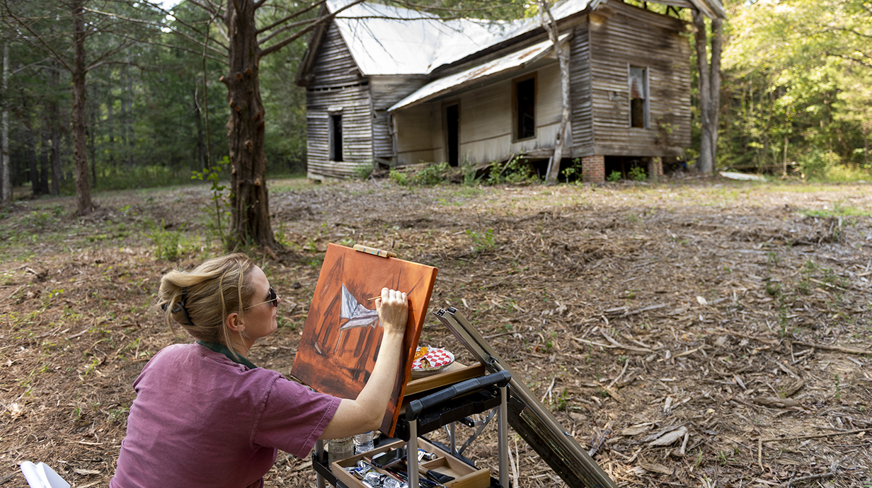 A woman sits outdoors at an easel, painting a nearby dilapidated building.