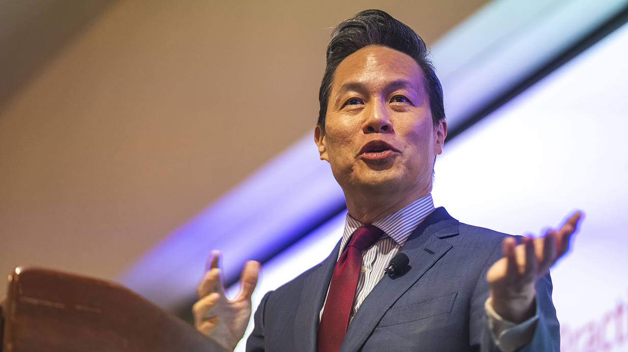 A man wearing a suit speaks in front of a projection screen.