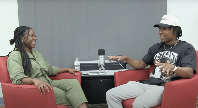 A young women and young man sit in easy chairs with a microphone on a tabletop between them.