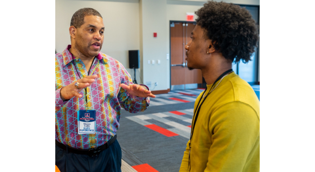 An older man talks with a young man standing in a conference hall.