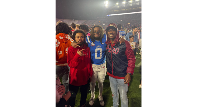 Three young men, one wearing a football uniform, stand in a hazy stadium with a large crowd milling about behind them at night.