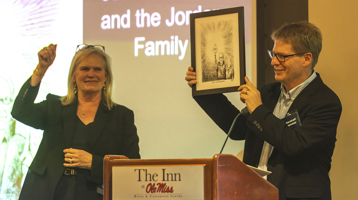 A man holds up a framed drawing on a stage in front of a projection screen as a woman raises her arm to cheer.