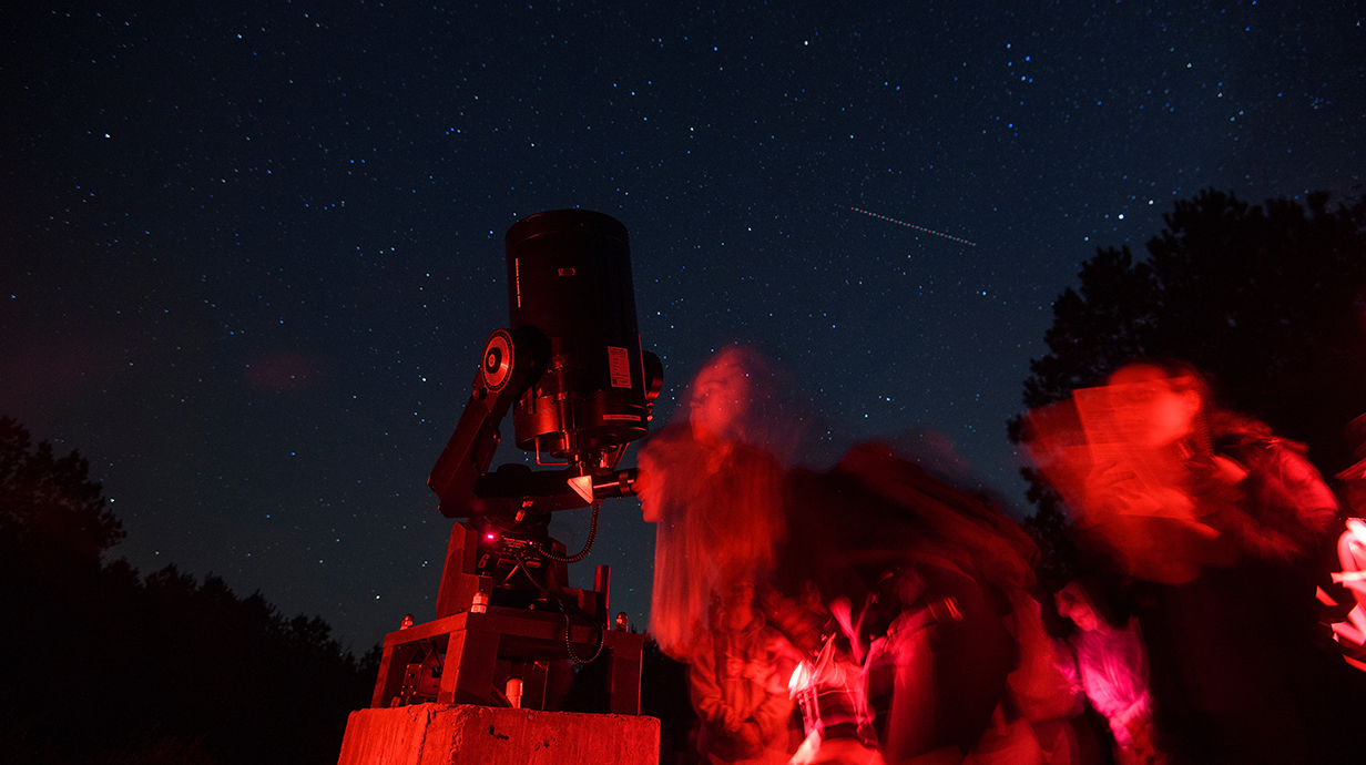 Several young people, lit by a red light, look through a telescope in front of a dary, star-filled sky.