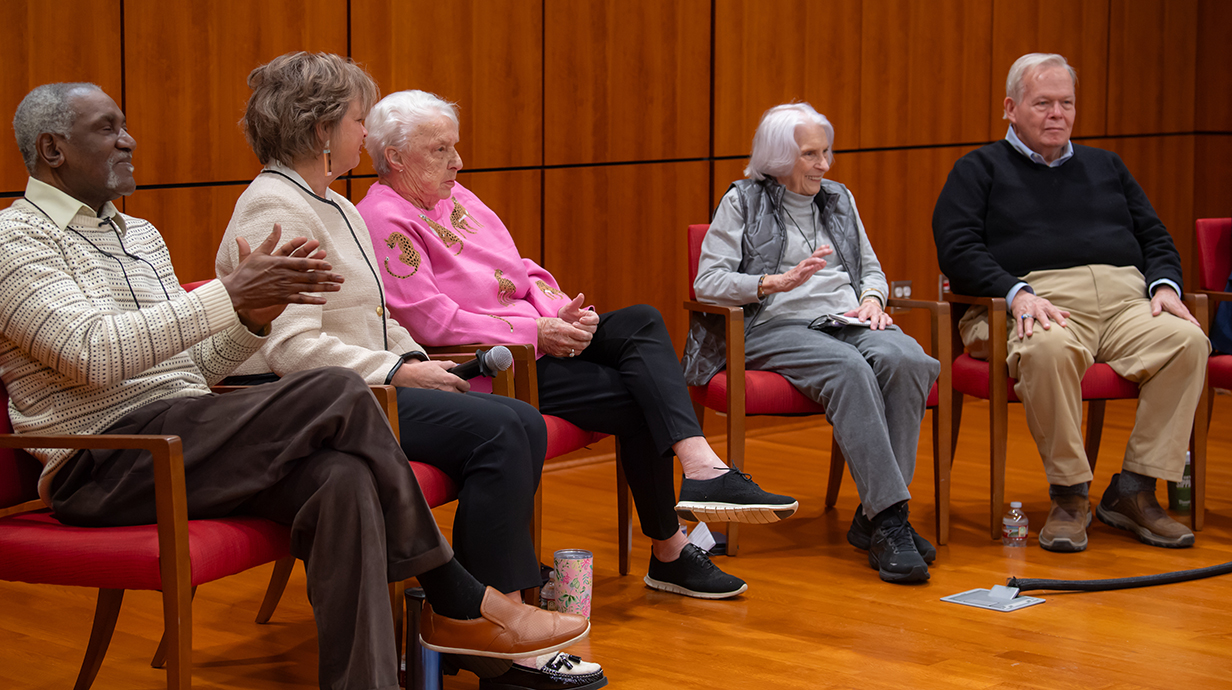 Three older women and two older men sit in chairs on a small raised platform in a conference room.