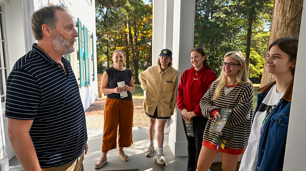 A man talks to a group of young people standing on the front porch of a large white building on a sunny day.