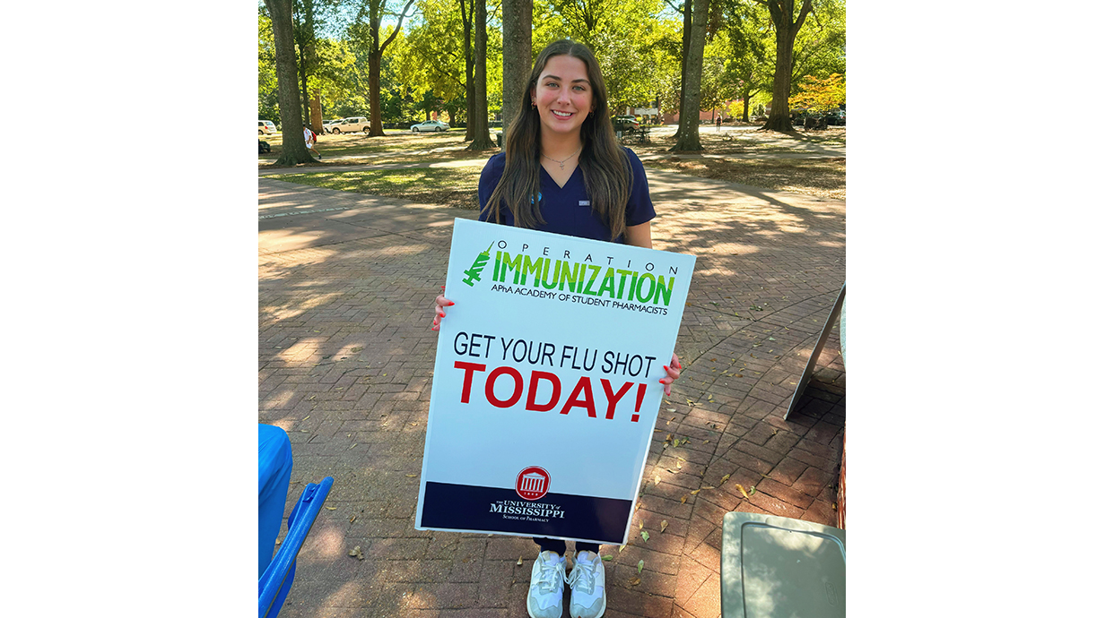 A young woman holds a large sign for Operation Immunization in an outdoor park.