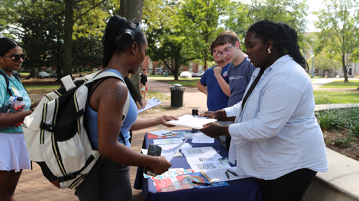 A young woman hands another young woman a clipboard and pen while several young people stand around an outdoor table lined with printed materials.