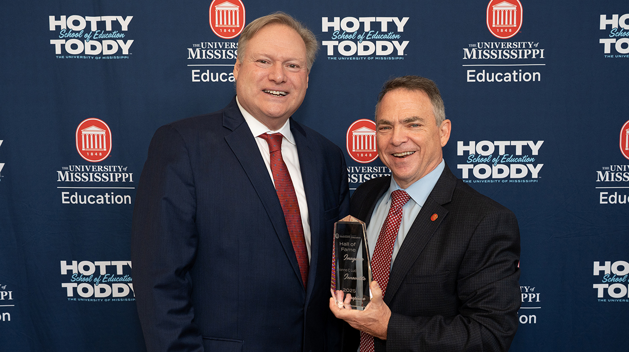 A man hands another man a trophy in front of a blue printed backdrop.