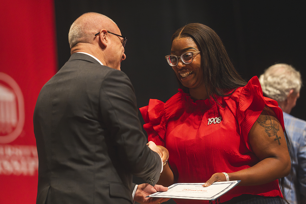 A woman in a red blouse with a "1908" brooch smiles as she receives a certificate and handshake from a bald man in a suit during a recognition event.