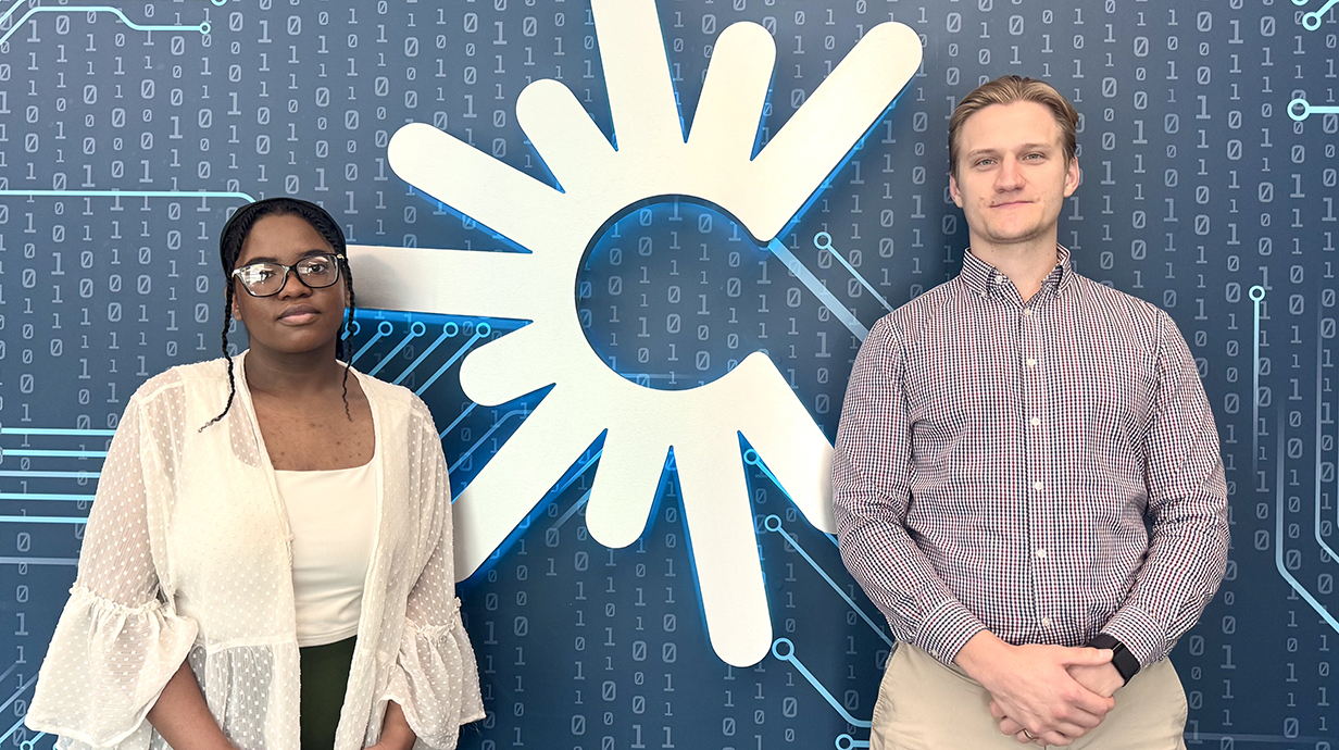 A young woman and a young man stand in front of a high-tech white logo on a dark blue background.