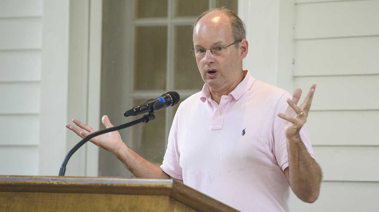 A man wearing a pink polo shirt gestures while speaking into a microphone in front of a white frame building.