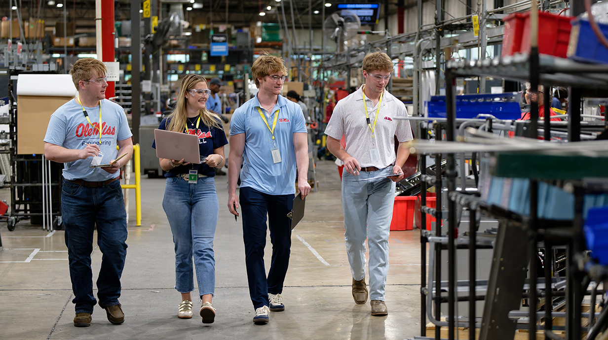 Three young men and a young woman, all wearing safety glasses and Ole Miss shirts and carrying clipboards, walk through a manufacturing plant.