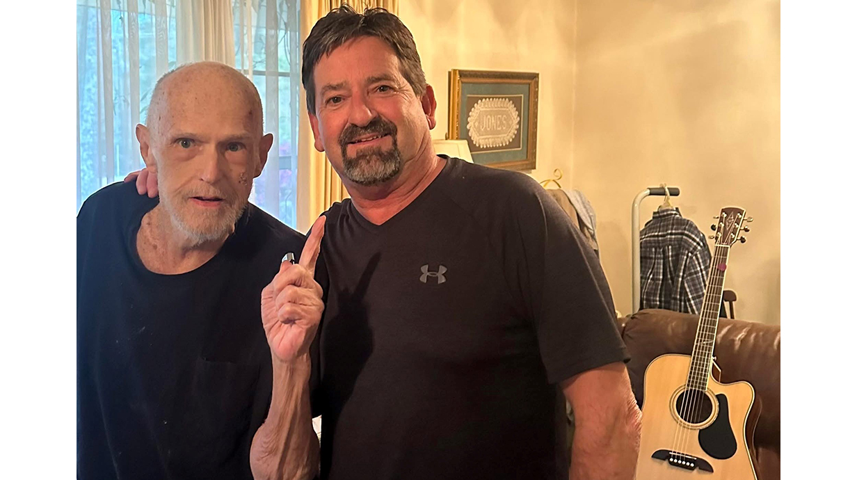 Two men have a photo taken together in a living room, with a guitar sitting on a couch behind them.