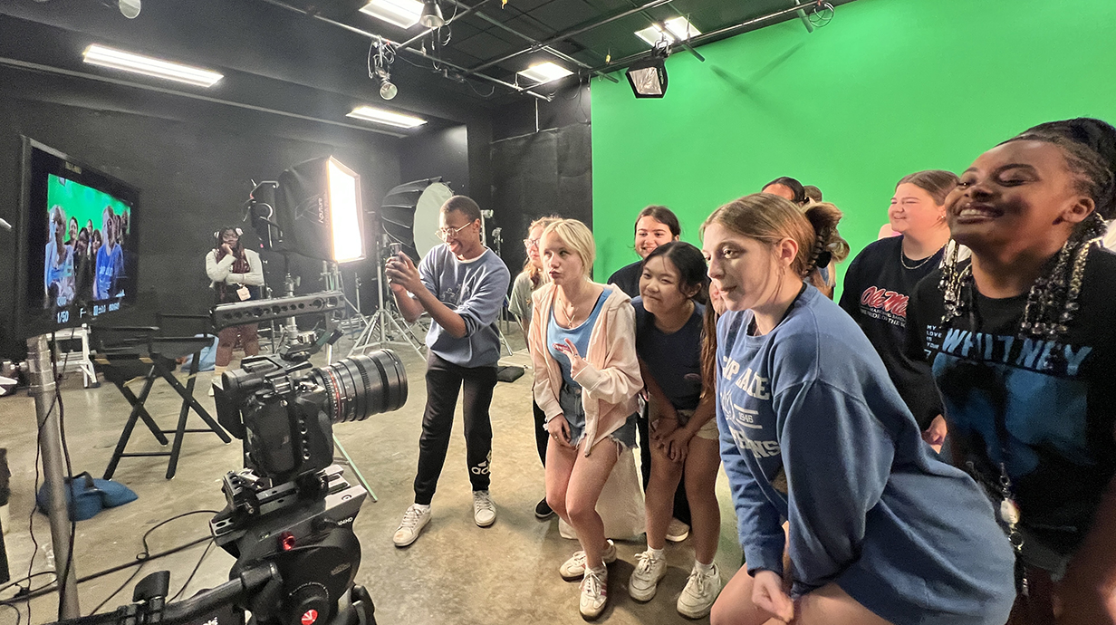A group of young people standing in front of a green backdrop look into a camera on a film soundstage.