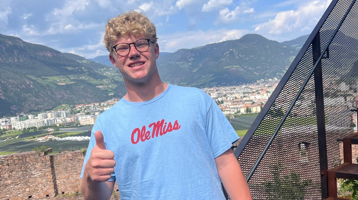 A young man wearing a blue T-shirt reading 'Ole Miss' stands on an outdoor stairway and gives a thumbs-up sign in front of a distant town skyline.