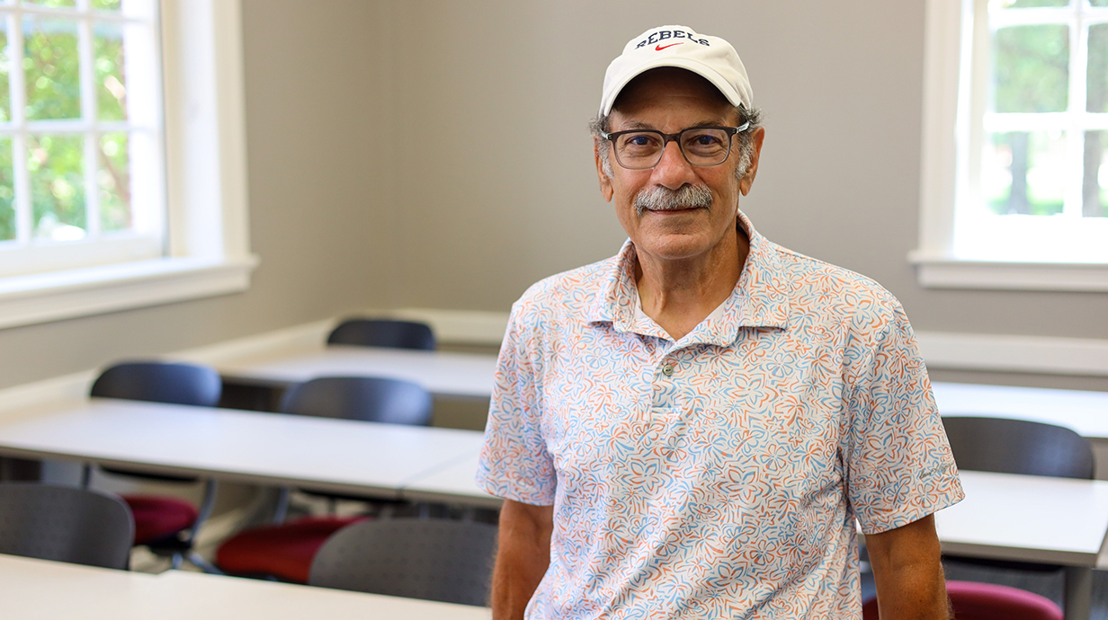 A man wearing a Rebels baseball cap stands in an empty classroom.