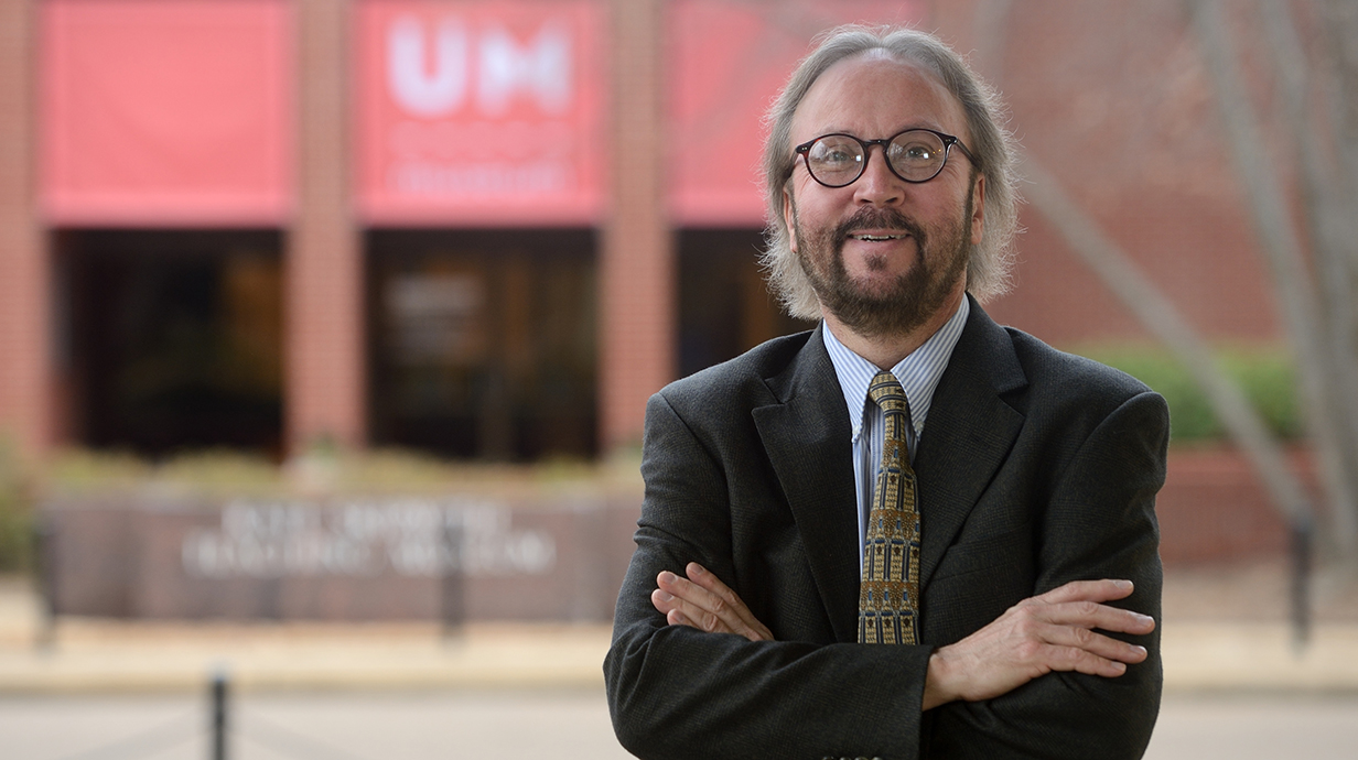 A man wearing a suit stands with arms crossed in front of a red brick building topped with a red banner reading 'UM Museum.'