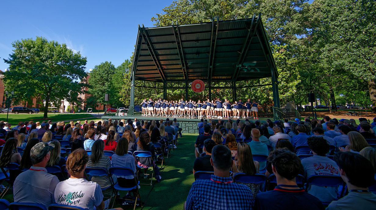 A large group of people sit in folding chairs in an outdoor park, watching a group of young people perform on an open-air stage.