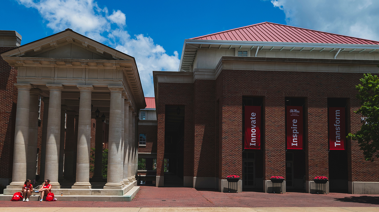 Two young women wearing ref T-shirts sit on the steps of a large white-columned porch in front of a tall red brick building with a brick courtyard surrounding it.