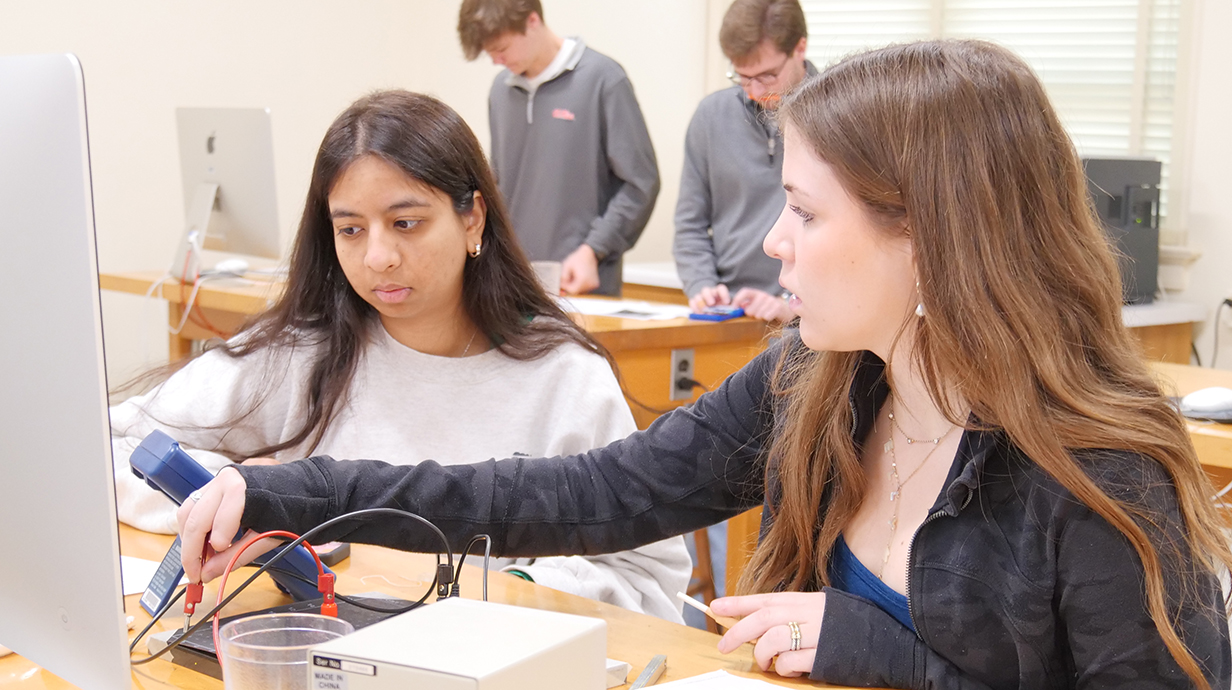 Two young women use an electronic device to complete a physics assignment in a classroom.