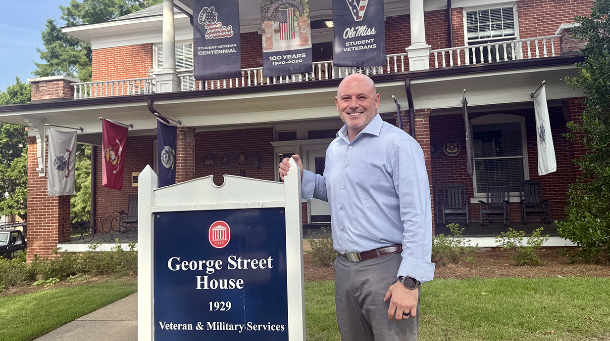 A man stands in front of a two-story brick house beside a sign reading 'George Street House.'