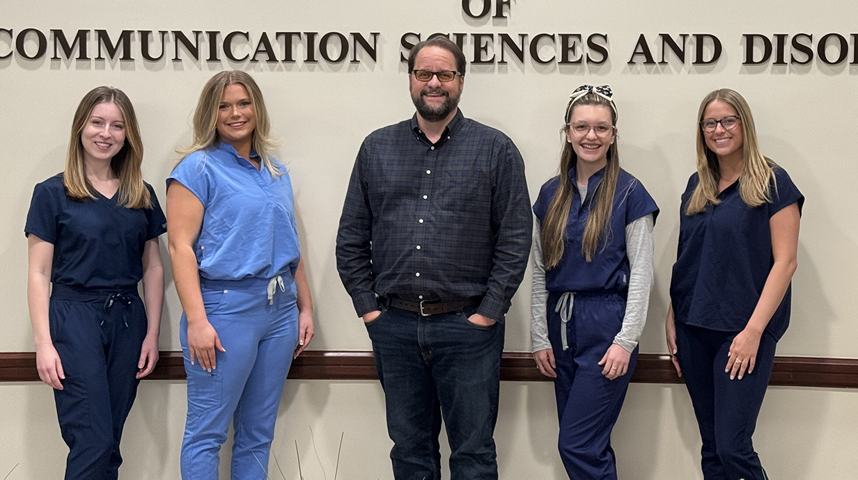 A man and four young women stand in front of a wall sign reading 'Department of Communication Sciences and Disorders.'