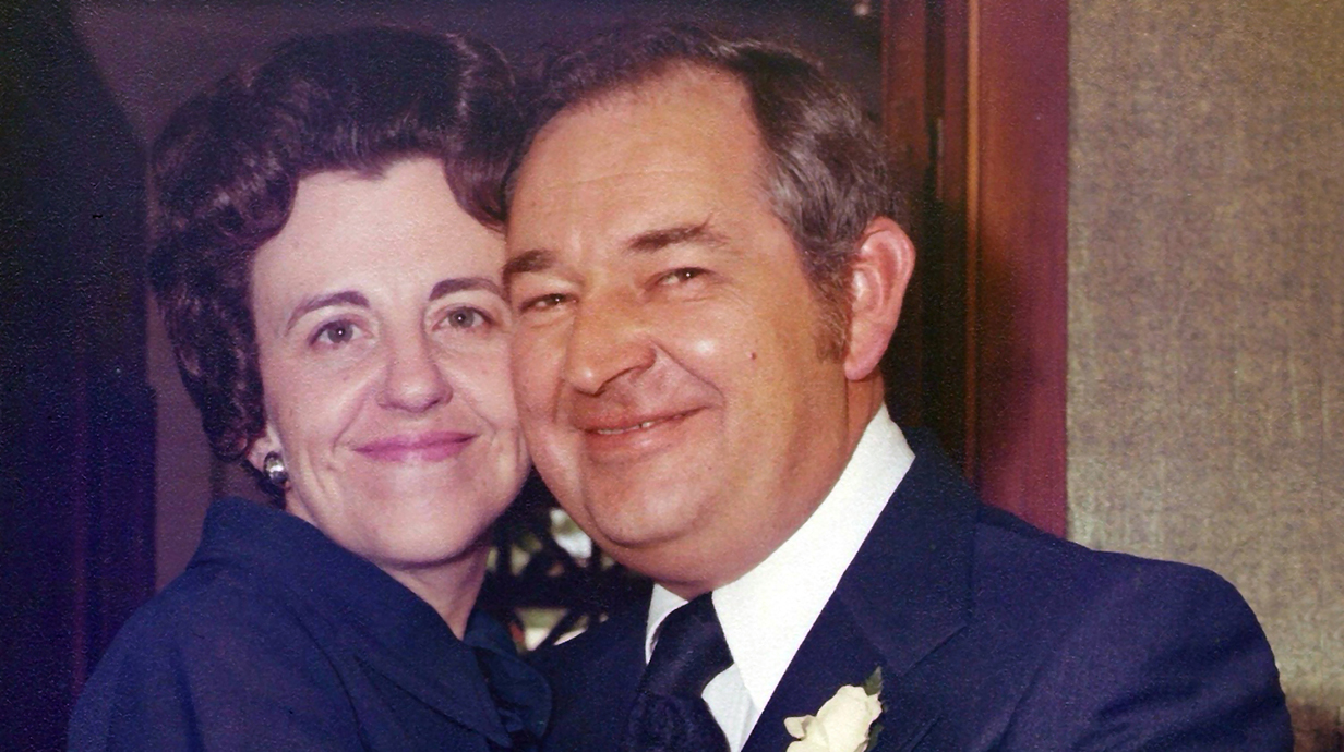 A man and woman, both wearing dress clothing, stand cheek-to-cheek for a portrait.