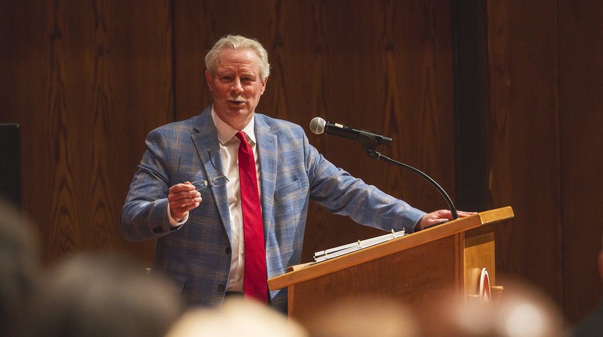 A man wearing a blue suit and a red necktie speaks to a crowd from a podium.
