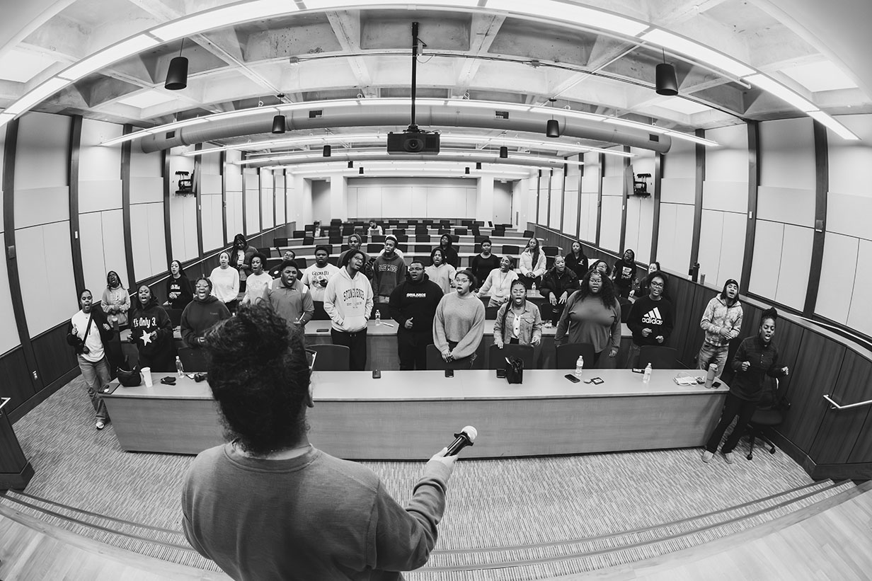 Wide-angle view of more than 40 University of Mississippi Gospel Choir members gathered in rows of chairs during rehearsal in the Student Union Auditorium, with the director standing at the front.