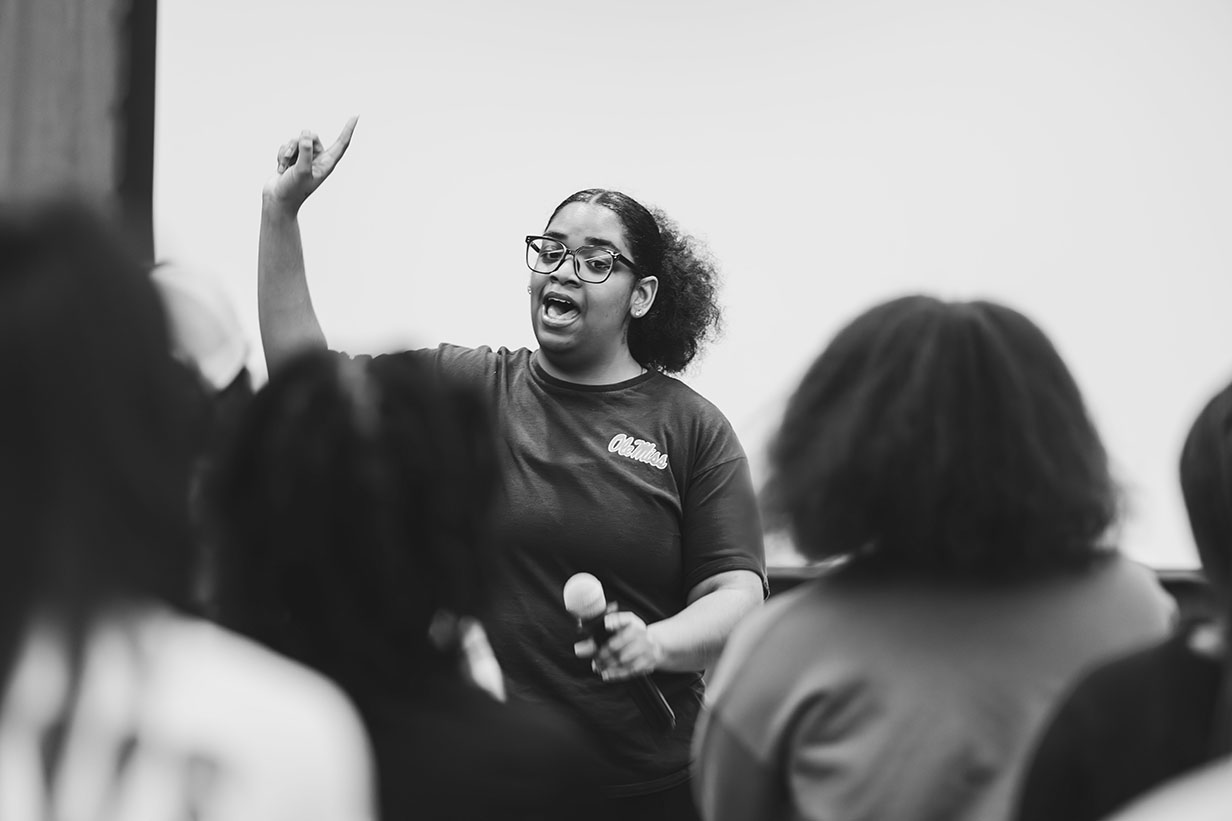 A young woman holds up her hand while talking to a group of people in an auditorium.