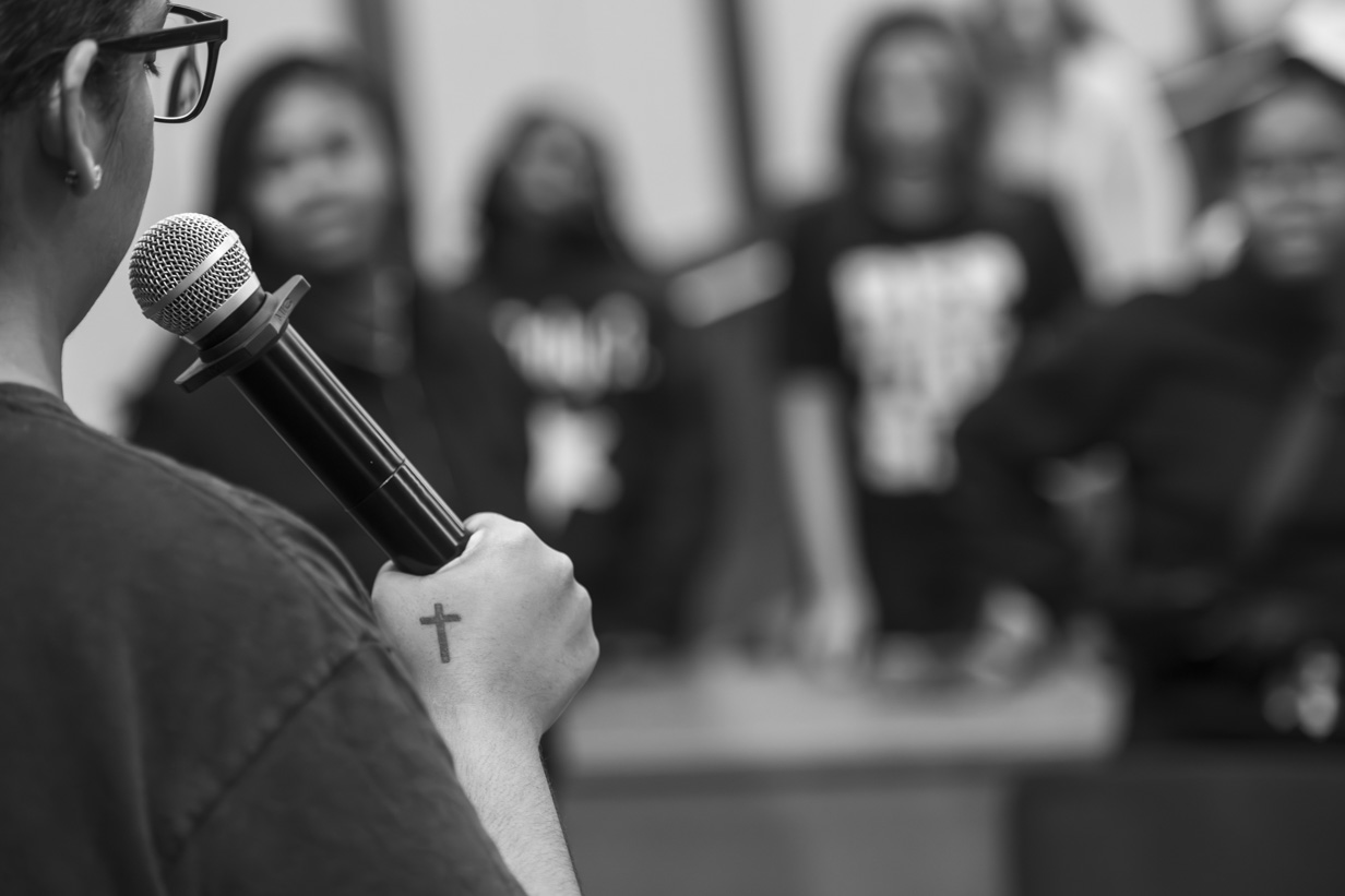 Close-up of choir director Honesty Shaw holding a microphone like a conductor's baton while directing the choir, with visible tattoos on her arms.
