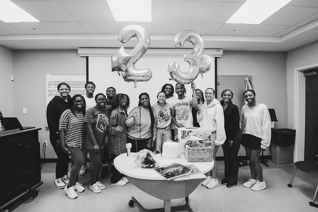 Group of Gospel Choir members celebrating around a table with birthday cake and decorations for director Honesty Shaw's surprise 23rd birthday party.