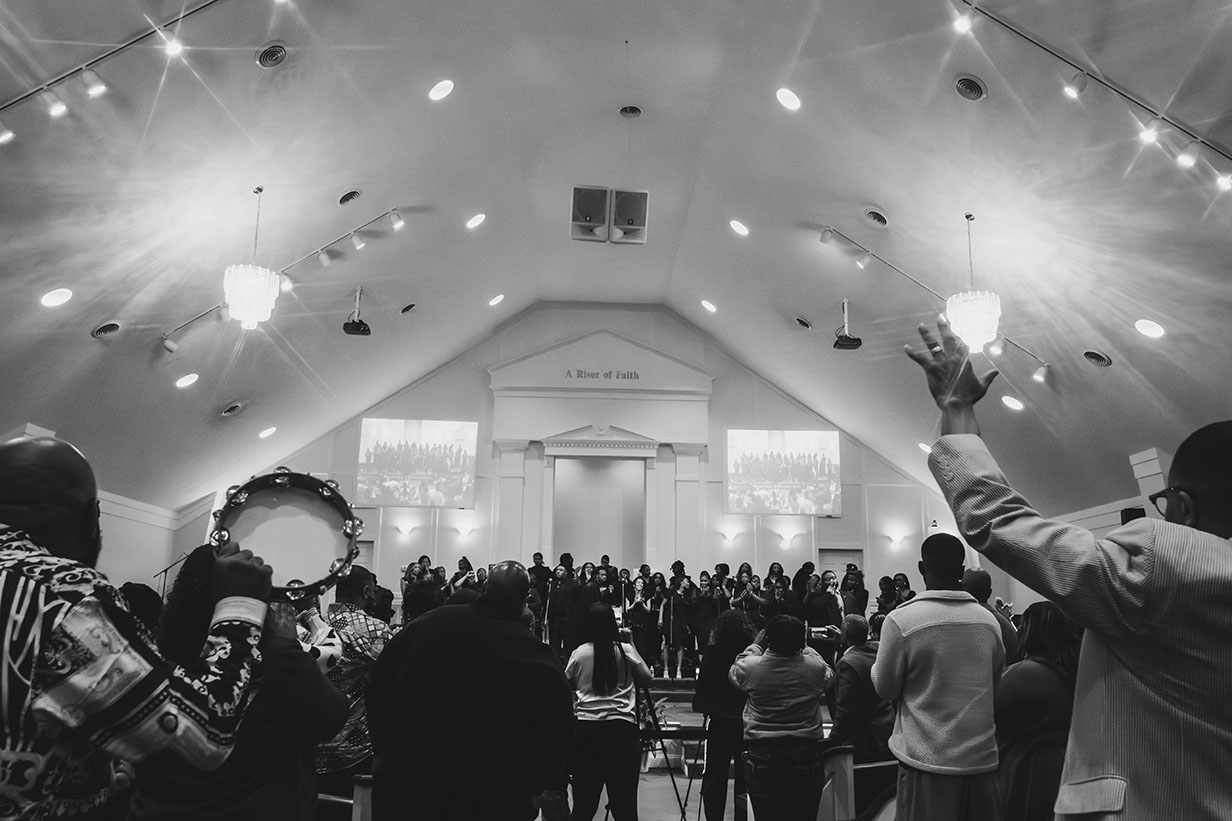 Concert audience members with raised hands and tambourines worshipping during the Gospel Choir's performance, with stage lights visible in the background.