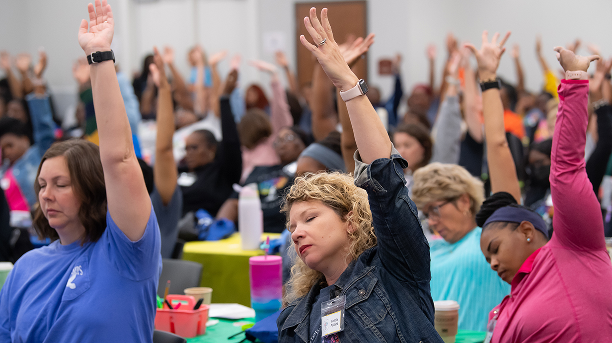 A large group of women practice a yoga pose with arms upraised and eyes closed.