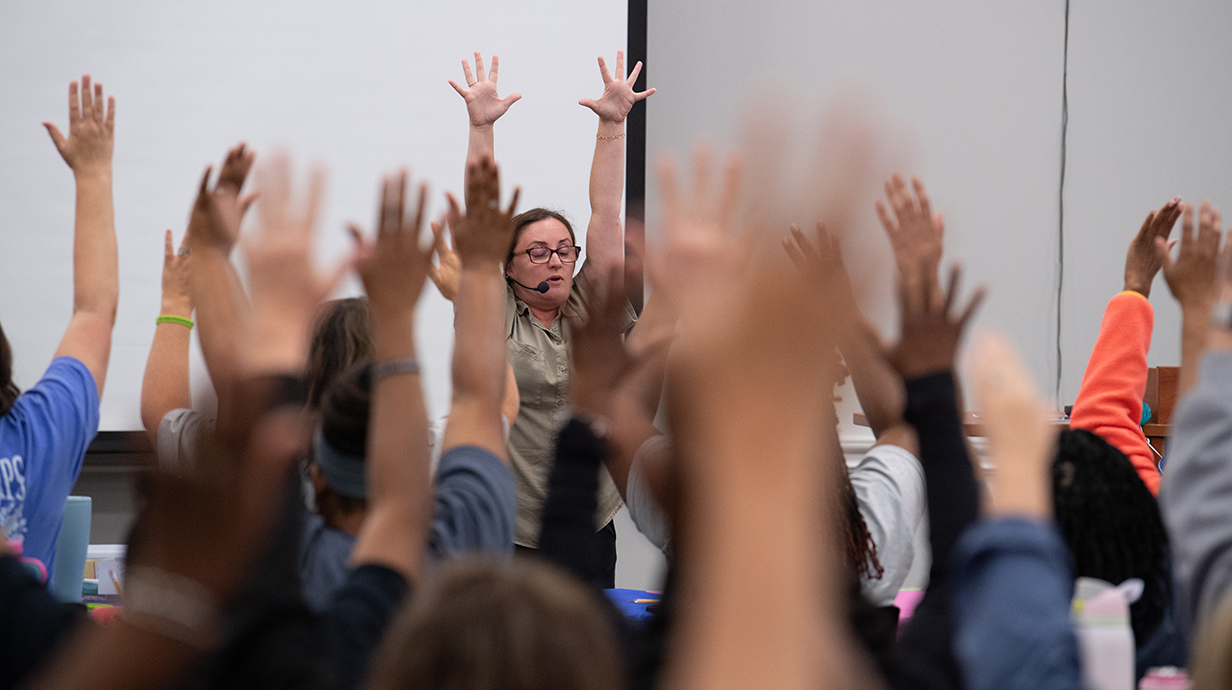 A woman wearing a microphone headset leads a group of women in a yoga pose.