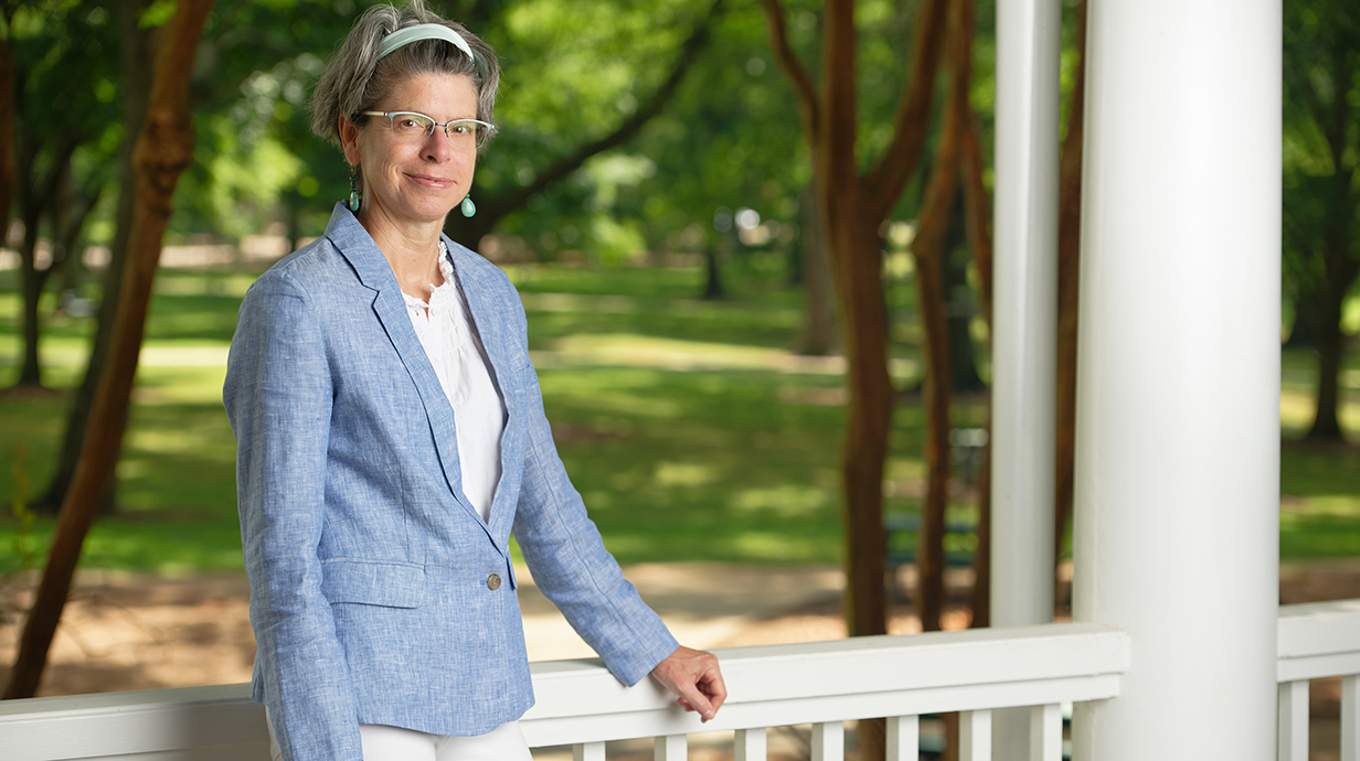 A woman stands at the railing of a covered porch overlooking a wooded park.