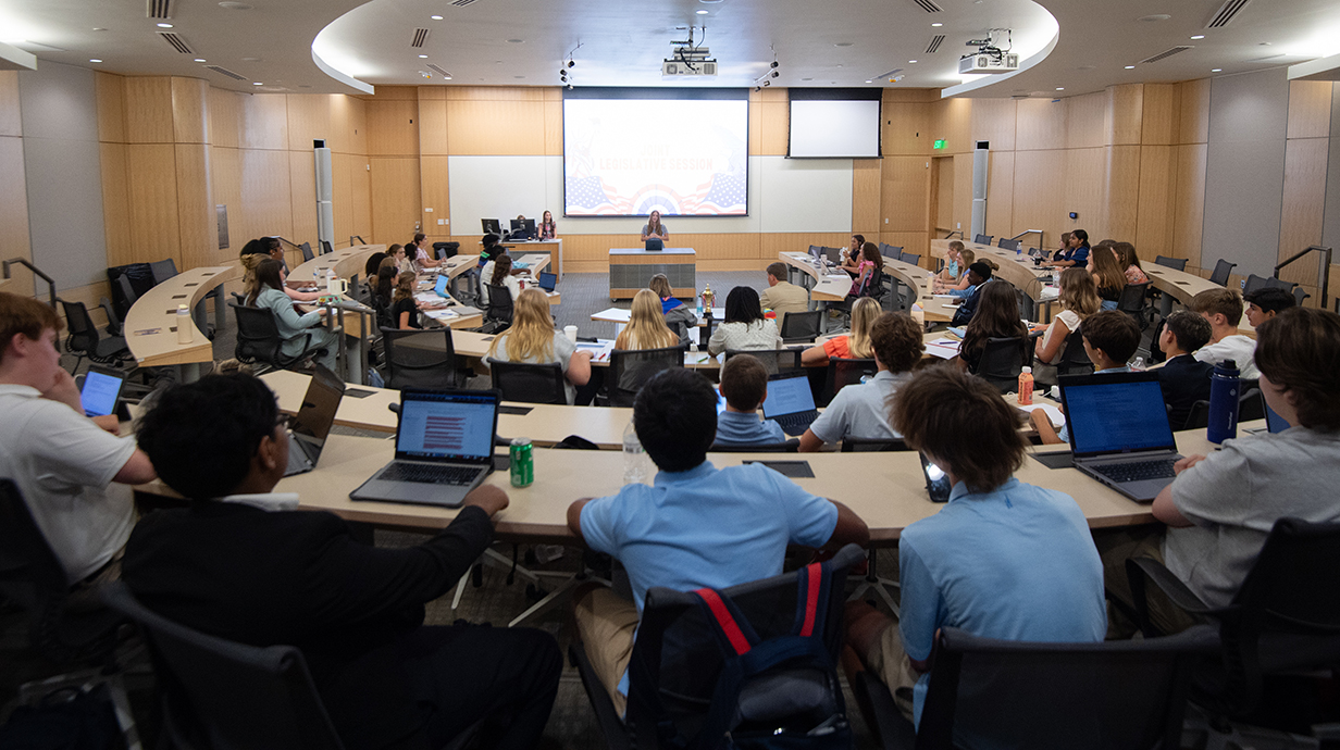 A young woman makes a presentation at the front of a large curved classroom.