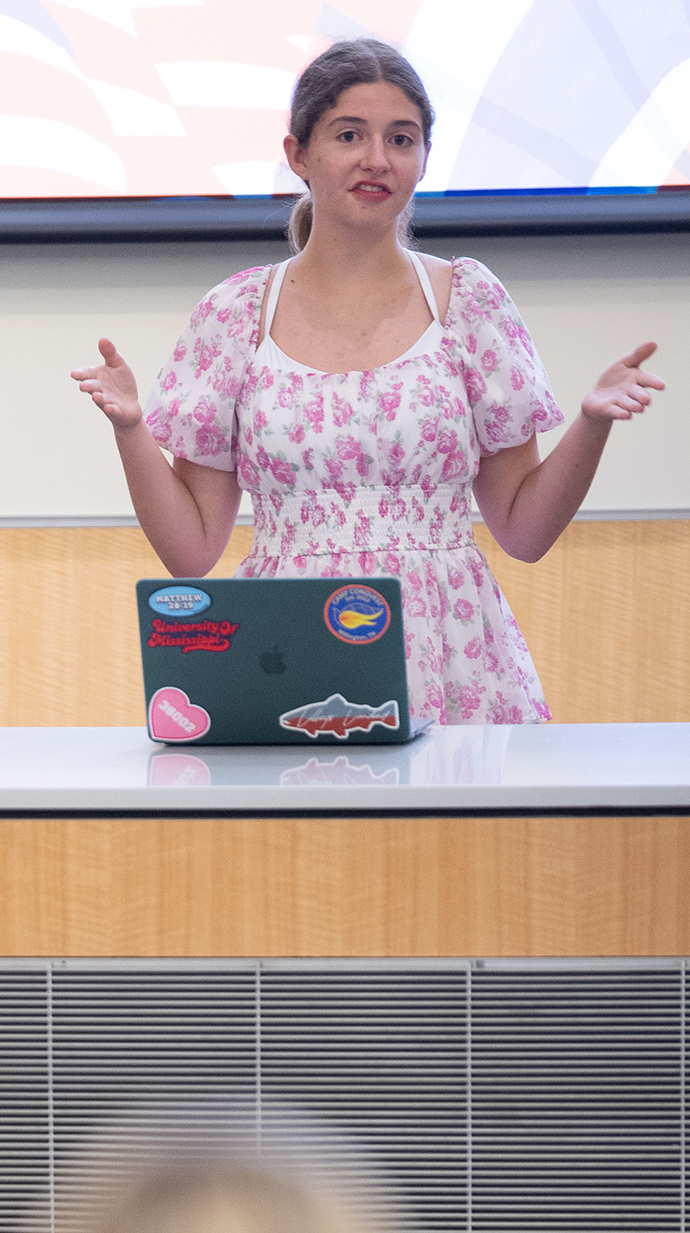 A young woman makes a presentation from a lectern in front of a projection screen.