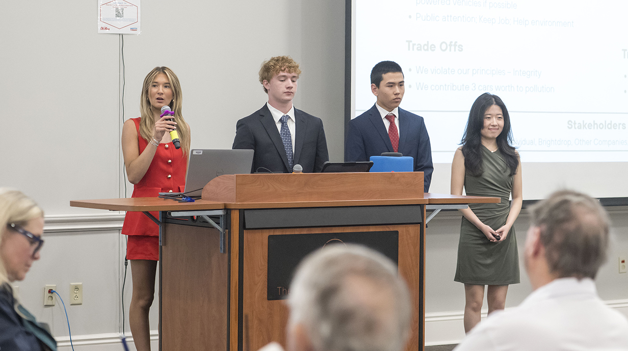 Two young women and two young men, all wearing business attire, speak to a crowd from a lectern in front of a projection screen.