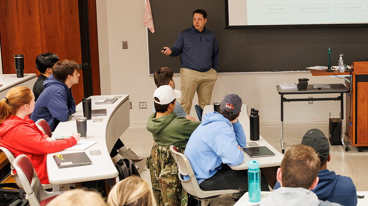 A man standing at a blackboard talks to students in a tiered auditorium.